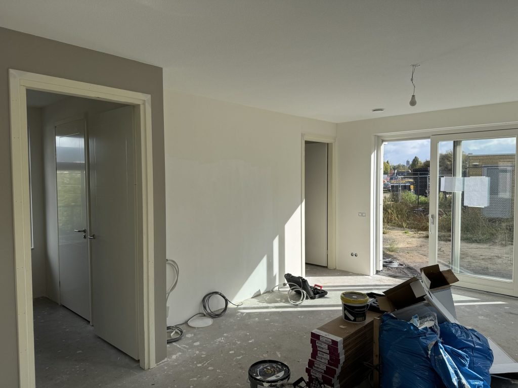 Nieuwe woonkamer tijdens de afwerking, met verfemmers en dozen op de betonvloer en tuindeuren naar een bouwterrein