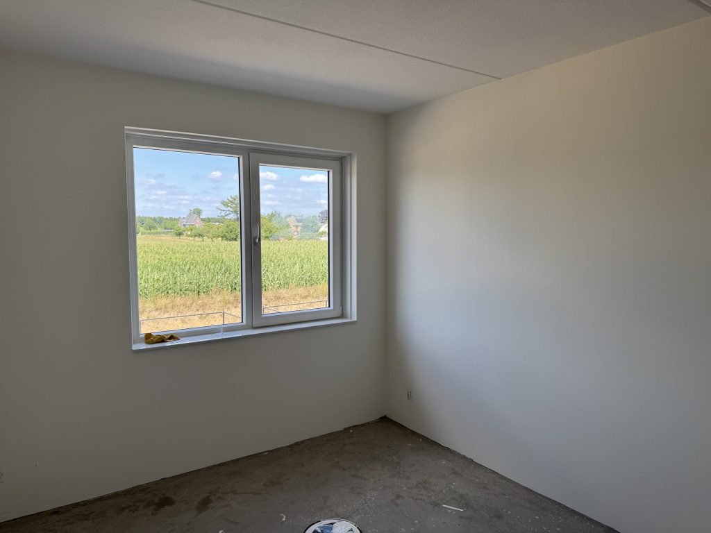 Lege nieuwbouwkamer met strak afgewerkte lichte wanden en een raam dat uitkijkt over een maisveld