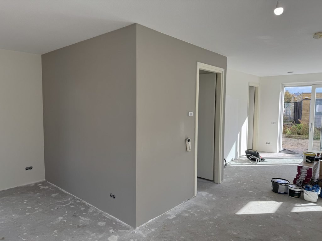 Nieuwbouwwoonkamer met een pas afgewerkte grijze wand rond de meterkast en verfemmers op de vloer