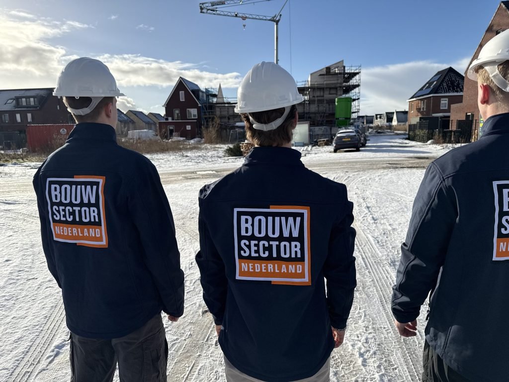 Drie vakmensen van Bouwsector Nederland met witte helm op een besneeuwde straat met woningen in aanbouw
